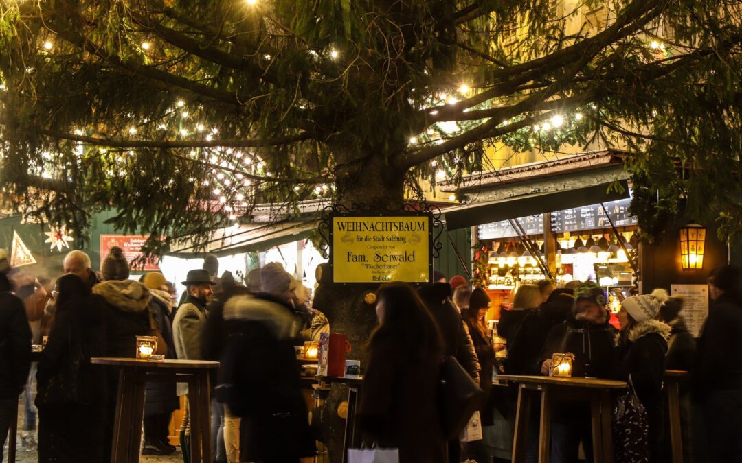 Besucher an Stehtischen unter dem beleuchteten Christbaum am Weihnachtsmarkt Mirabellplatz am Abend