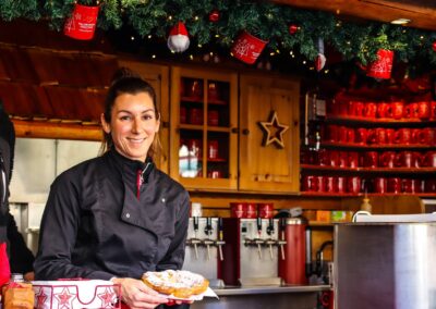 Lächelnde Mitarbeiterin der Christl-Alm hält einen Bauernkrapfen, dahinter Regale mit roten Häferln