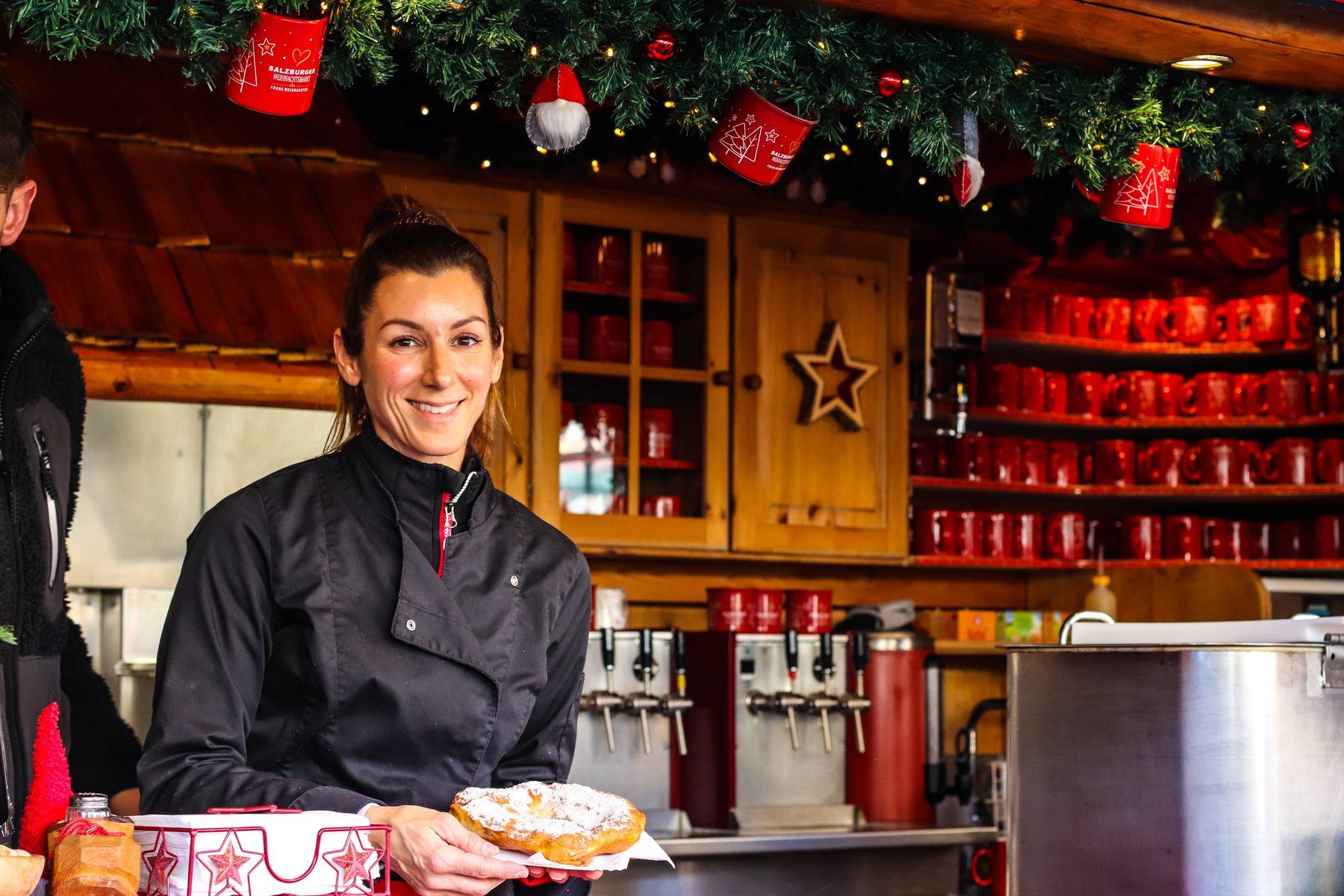 Lächelnde Mitarbeiterin der Christl-Alm hält einen Bauernkrapfen, dahinter Regale mit roten Häferln Lächelnde Mitarbeiterin der Christl-Alm hält einen Bauernkrapfen, dahinter Regale mit roten Häferln