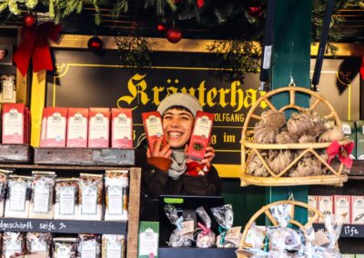 Standlerin hält zwei Packungen Früchtetee unter dem Schild Kräuterhaus in der Hütte am Weihnachtsmarkt Mirabellplatz