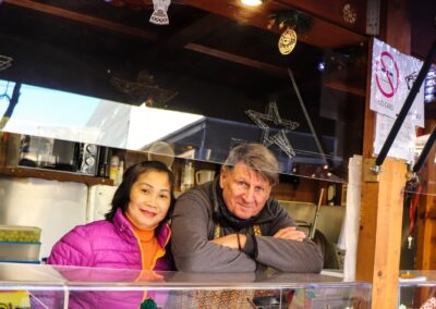 Zwei Standler lehnen lächelnd an der Glasvitrine der Hütte von Ludwig Glier am Weihnachtsmarkt Mirabellplatz