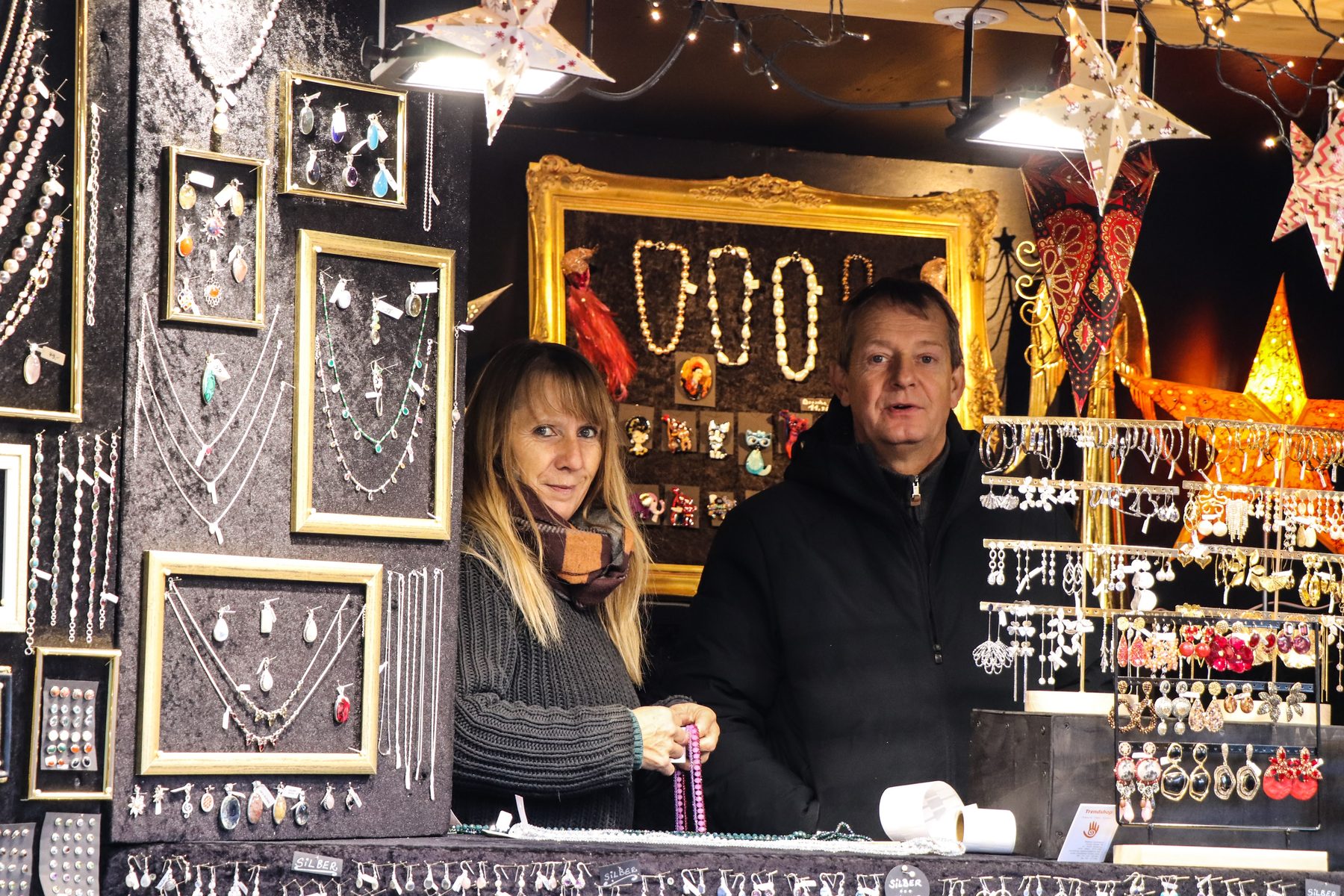 Standlerin und Standler in der Schmuckhütte von Markus Germayer zwischen gerahmten Ketten am Weihnachtsmarkt Mirabellplatz